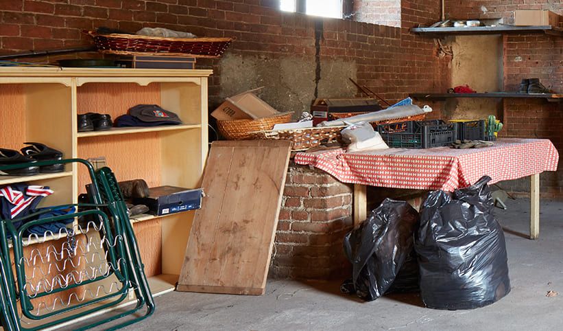 Überfüllter Kellerraum mit Regalen, einem mit verschiedenen Gegenständen bedeckten Tisch, Müllsäcken und einem an die Wand gelehnten Holzbrett
