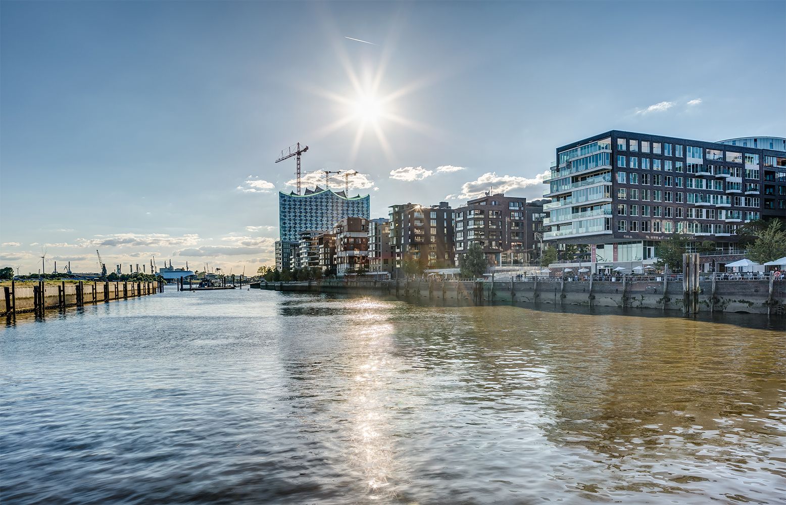 Die Sonne scheint über dem Hafen, beleuchtet moderne Gebäude und Spiegelungen auf dem Wasser, mit Kränen im Hintergrund